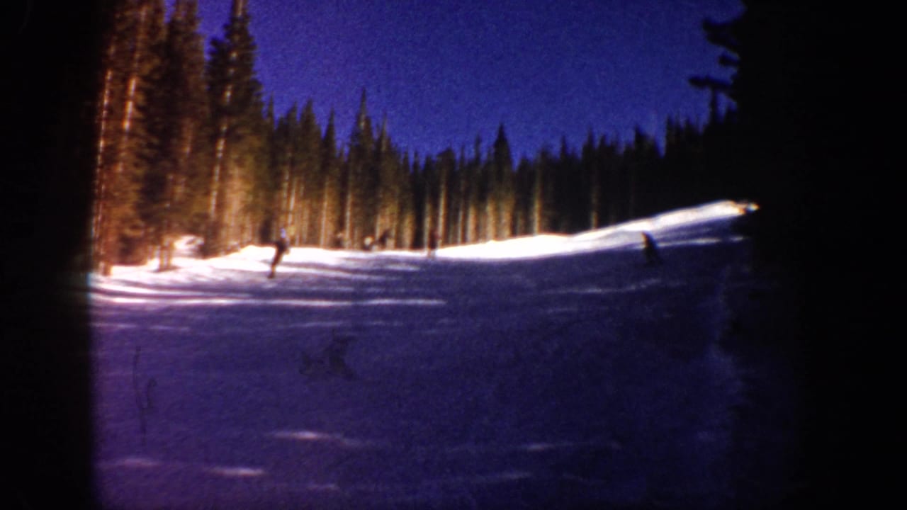 Skiers on a dim snowy slope bordered by evergreen trees under a blue dusk sky.