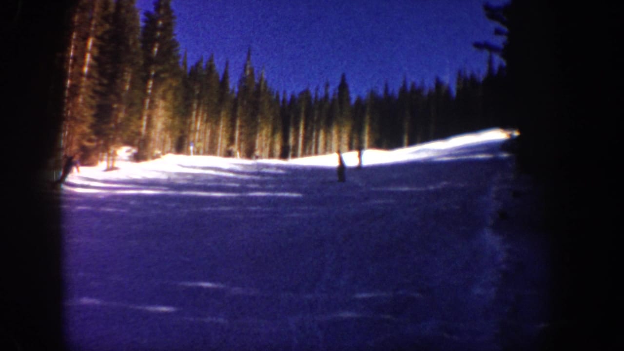 1961 Aspen ski-area footage shows small skier silhouettes on a blue-purple snowy run bordered by tall evergreens.