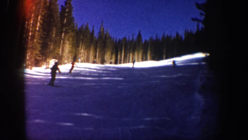 1961 Aspen 8mm frame with multiple skiers on a shadowed slope, framed by evergreens and bright snow at dusk.