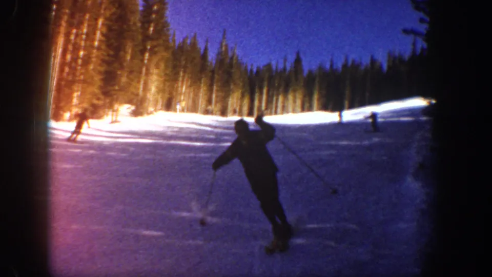 1961 Aspen home-movie frame of a foreground skier turning on a snowy slope, with distant skiers and evergreens behind.