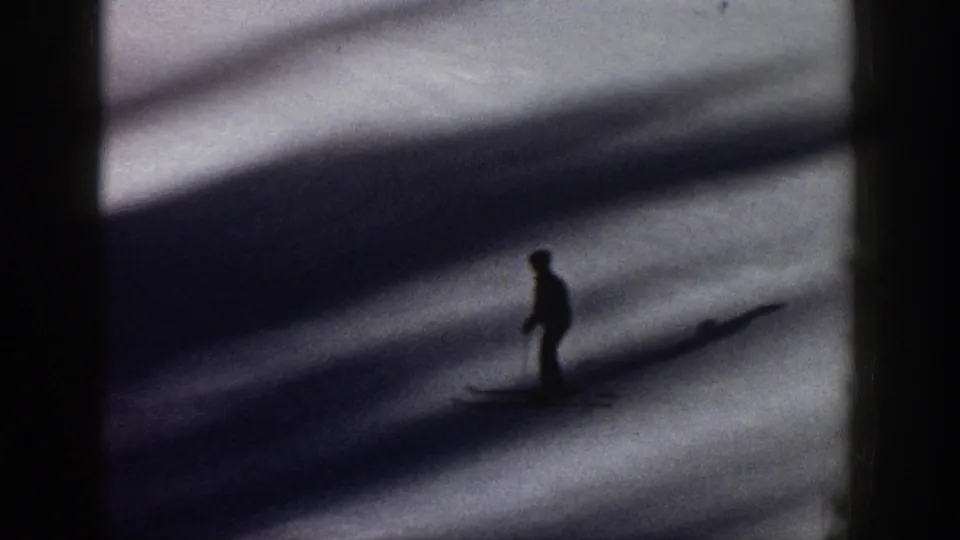 A silhouetted skier moves across shadowed snow in 1961 Aspen home-movie footage, with heavy contrast and soft 8mm grain.