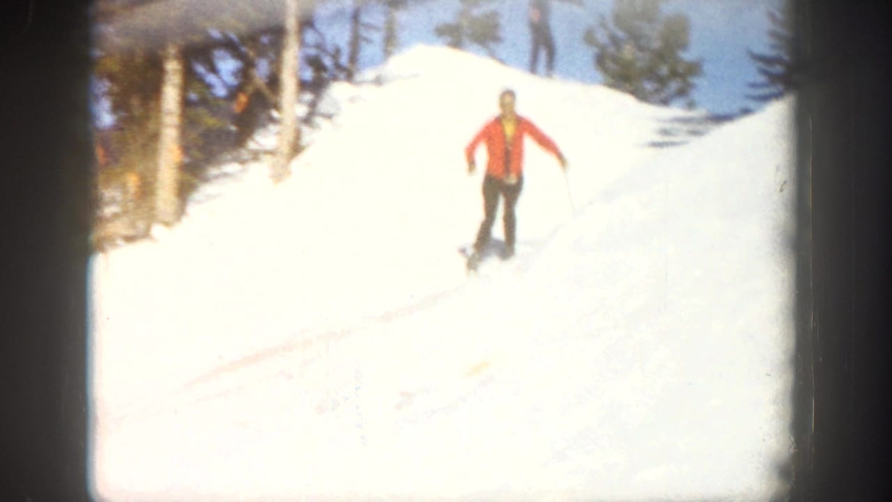 A skier in a red jacket and dark pants descends a bright Aspen snow slope with evergreen trees behind, filmed in soft 8mm color.