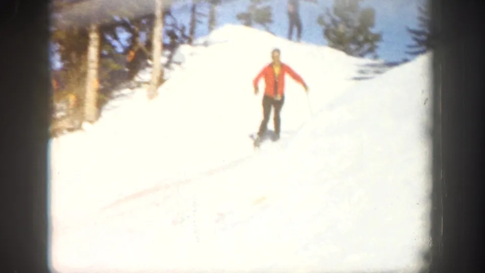 A skier in a red jacket and dark pants descends a bright Aspen snow slope with evergreen trees behind, filmed in soft 8mm color.