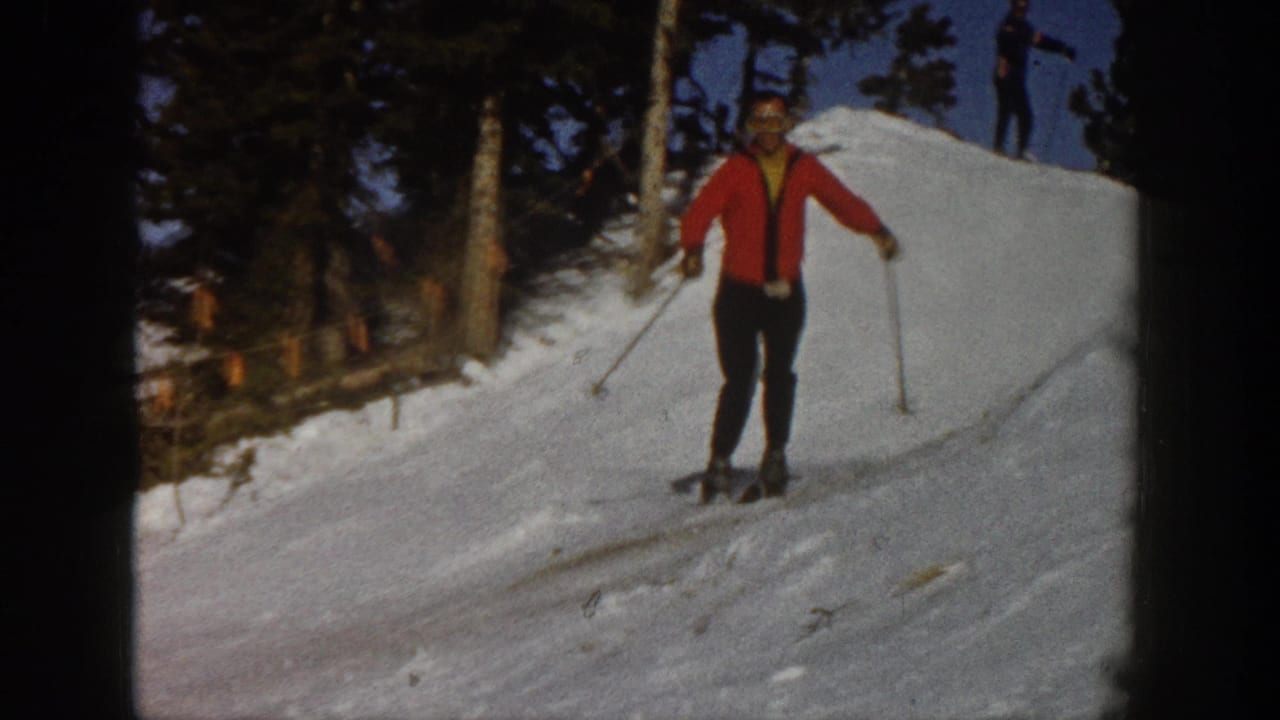 A red-jacketed skier approaches the camera on a snowy Aspen slope, with trees at the side and another skier visible on the ridge.