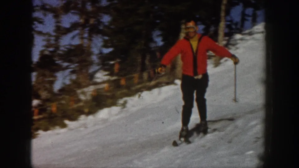 In the supplied 1961 Aspen footage, a skier in a red sweater and goggles approaches on a snowy, tree-lined slope.