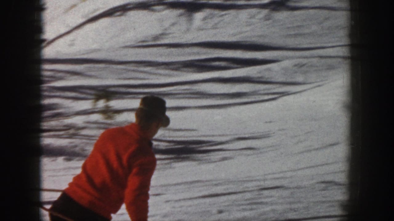 A skier in red turns across a snowy Aspen slope, seen from behind or side view with tree shadows across the snow.