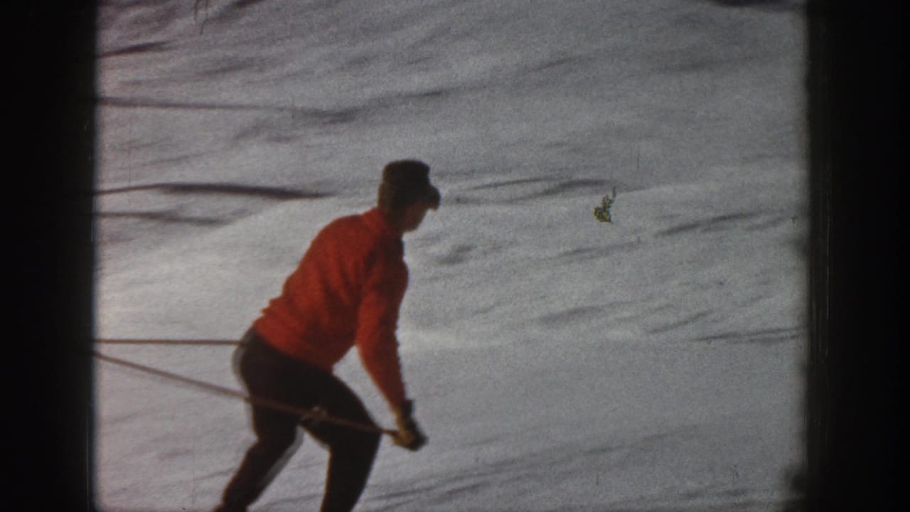 1961 Aspen home-movie frame showing a skier in a red jacket and dark pants moving downhill on snow.