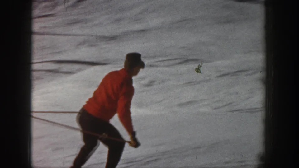 1961 Aspen home-movie frame showing a skier in a red jacket and dark pants moving downhill on snow.