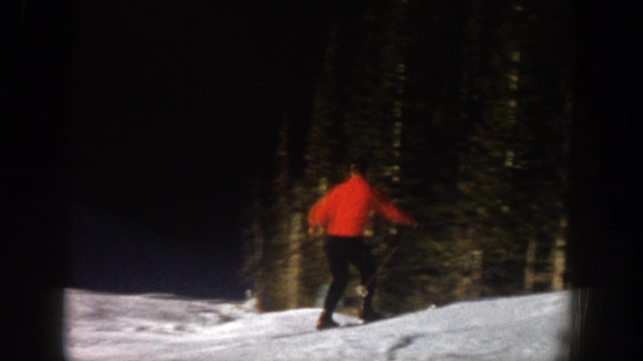 Home-movie frame from Aspen shows the skier moving downhill near evergreen trees in dimmer exposure.