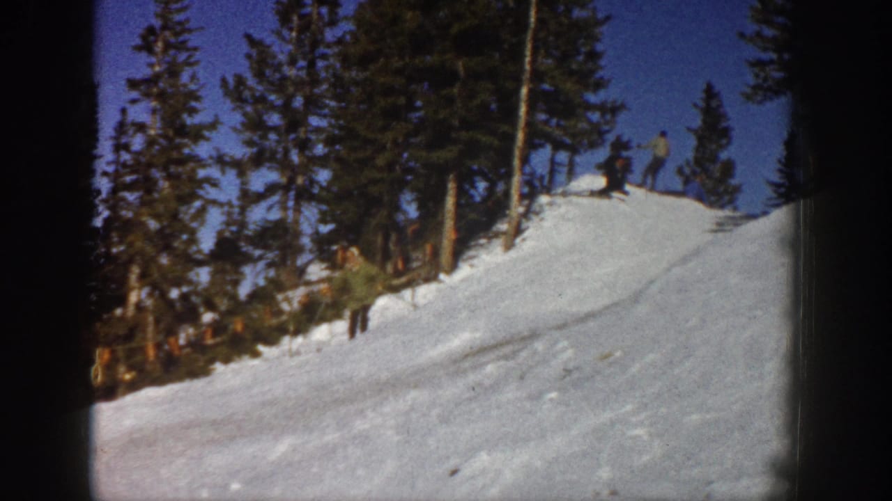Aspen 1961 frame showing a snowy slope, evergreen trees, blue sky, and distant skiers or observers.