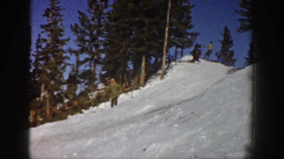 Aspen 1961 frame showing a snowy slope, evergreen trees, blue sky, and distant skiers or observers.