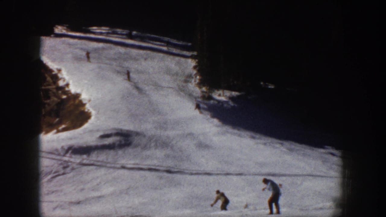 Aspen 1961 home-movie frame with one skier bending or adjusting gear while other skiers move downhill.