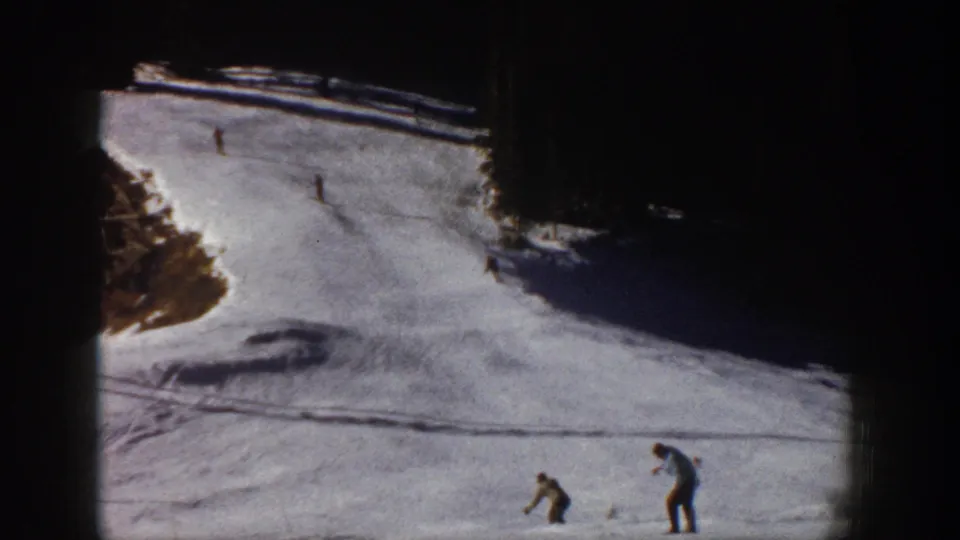 Aspen 1961 home-movie frame with one skier bending or adjusting gear while other skiers move downhill.