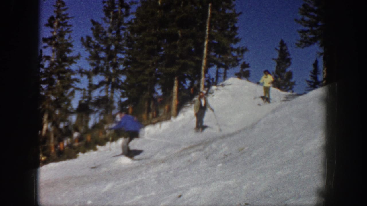 1961 Aspen home-movie frame showing skiers near an evergreen-lined snowy rise under a clear blue sky.