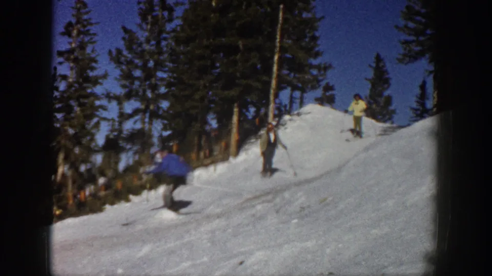 1961 Aspen home-movie frame showing skiers near an evergreen-lined snowy rise under a clear blue sky.