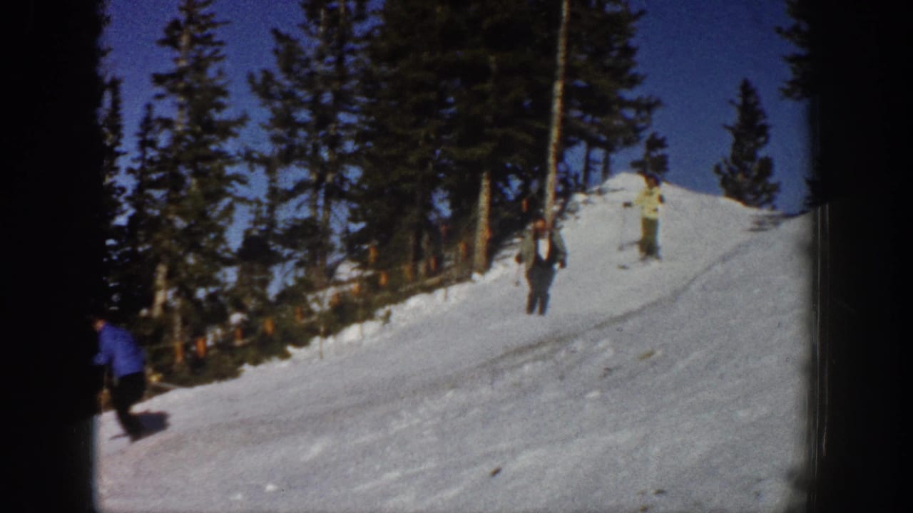 1961 Aspen home-movie frame showing several skiers spread across a snowy slope beneath evergreen trees and blue sky.