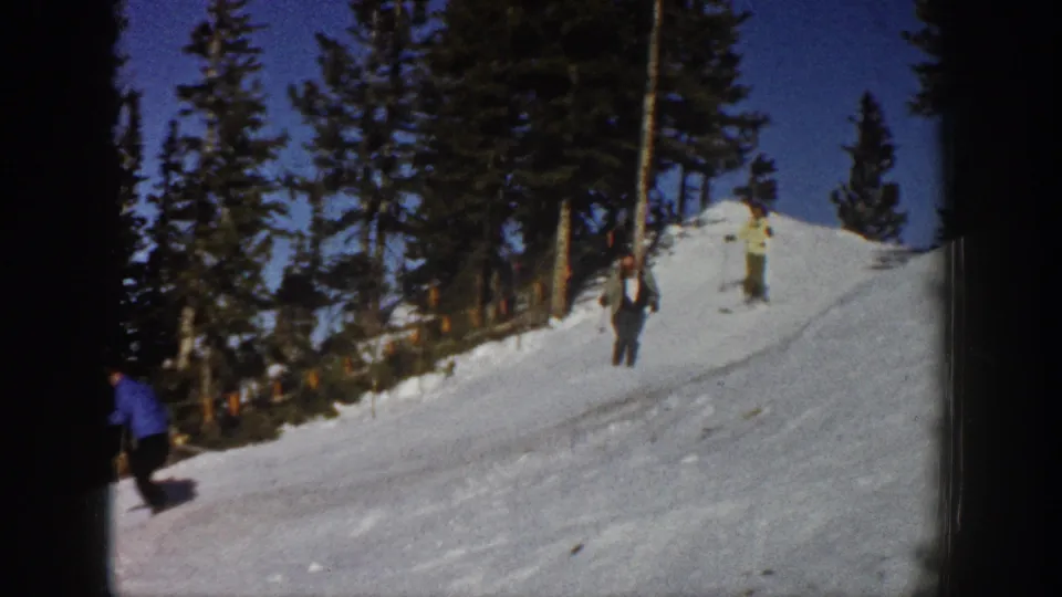 1961 Aspen home-movie frame showing several skiers spread across a snowy slope beneath evergreen trees and blue sky.