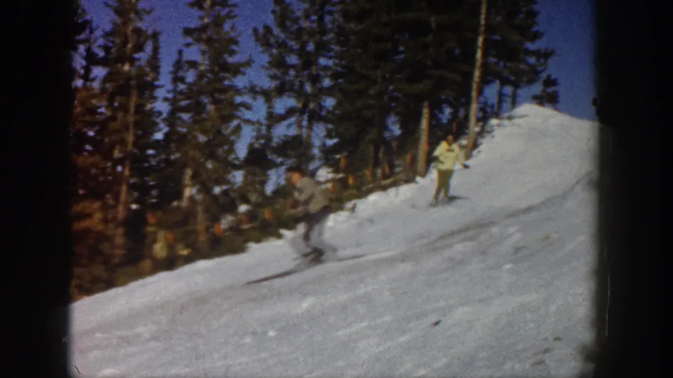 A skier in dark clothing moves downhill on a snowy Aspen slope, with another skier and evergreen trees in the background.