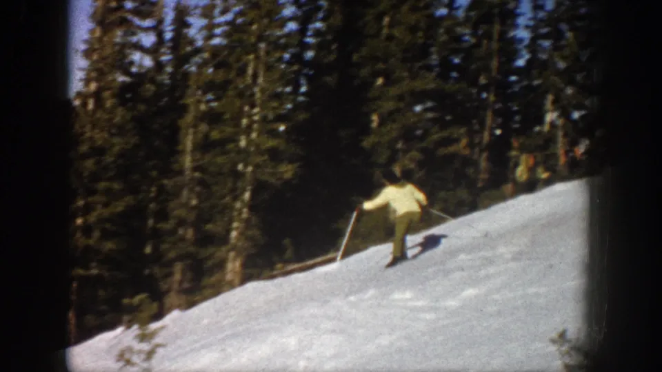 In 1961 Aspen, Colorado, a woman in a light jacket practices skiing on a snow-covered slope bordered by evergreens.