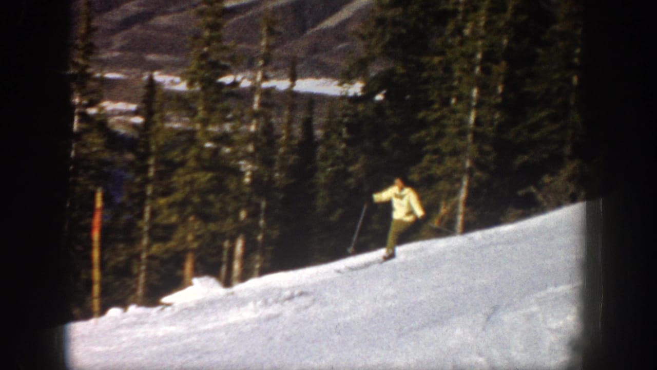 The woman skier moves along a snowy Aspen hillside, holding poles as evergreens and mountain terrain fill the background.