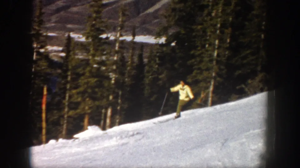 The woman skier moves along a snowy Aspen hillside, holding poles as evergreens and mountain terrain fill the background.