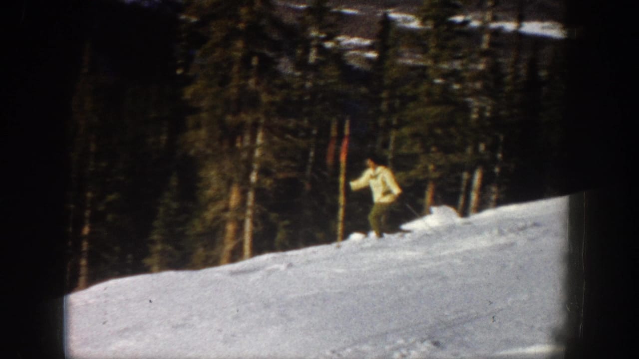 The woman slows or steadies herself on the Aspen snow slope, framed by dense evergreens and the soft texture of 8mm film.