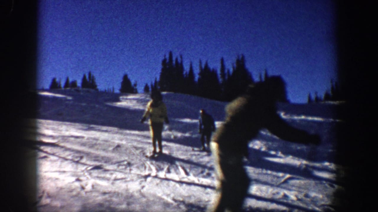 Home-movie frame from 1961 Aspen showing a skier crossing near the camera with two figures on the snowy slope beyond.
