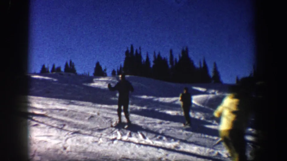 1961 Aspen home-movie frame showing several skiers on a snowy slope with dark evergreen trees and strong winter light.