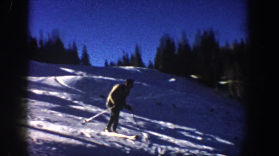 1961 Aspen winter frame showing a skier in profile crossing a bright slope with ski tracks and evergreens behind.