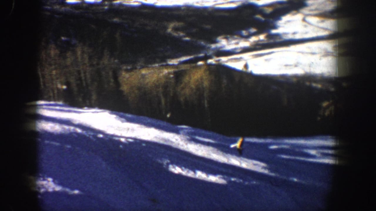1961 Aspen, Colorado home-movie frame showing skiers on a bright snowy slope with pine trees and distant terrain beyond.
