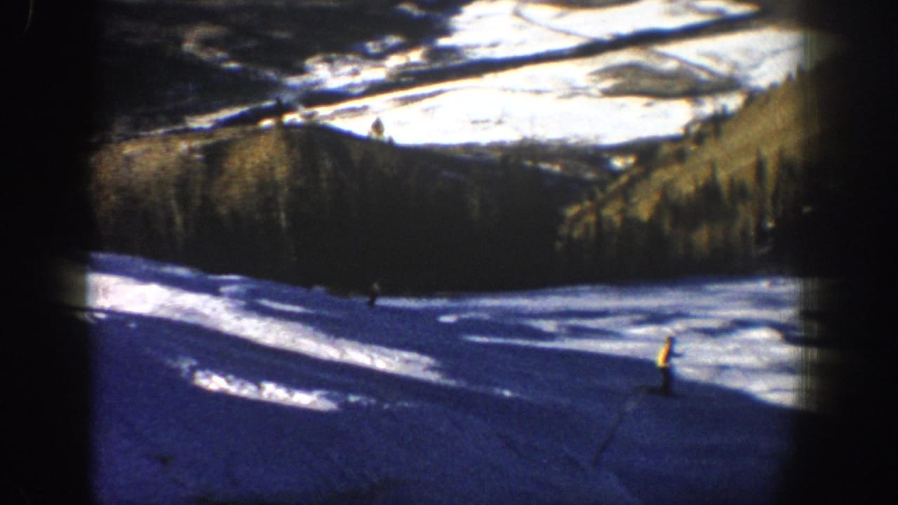 Two distant skiers cross a bright slope with trees and snowy ground behind them.