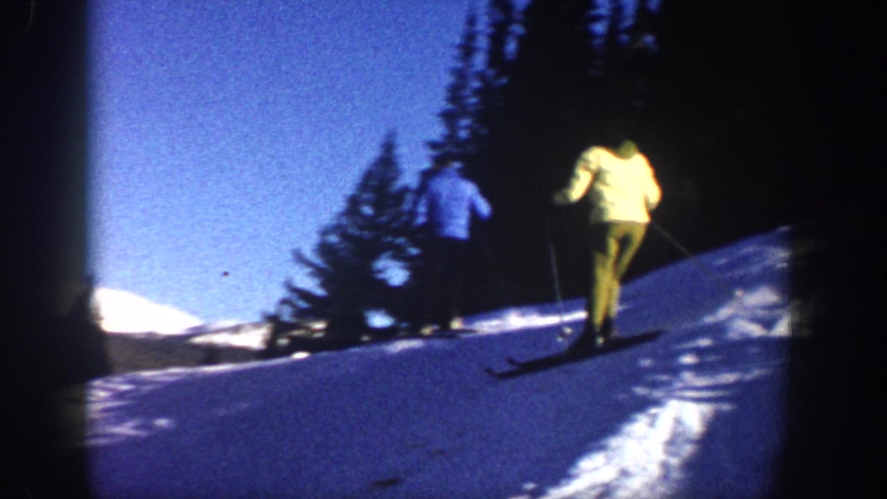 1961 Aspen frame showing two distant skiers crossing a bright snowy slope with evergreen trees and mountains beyond.