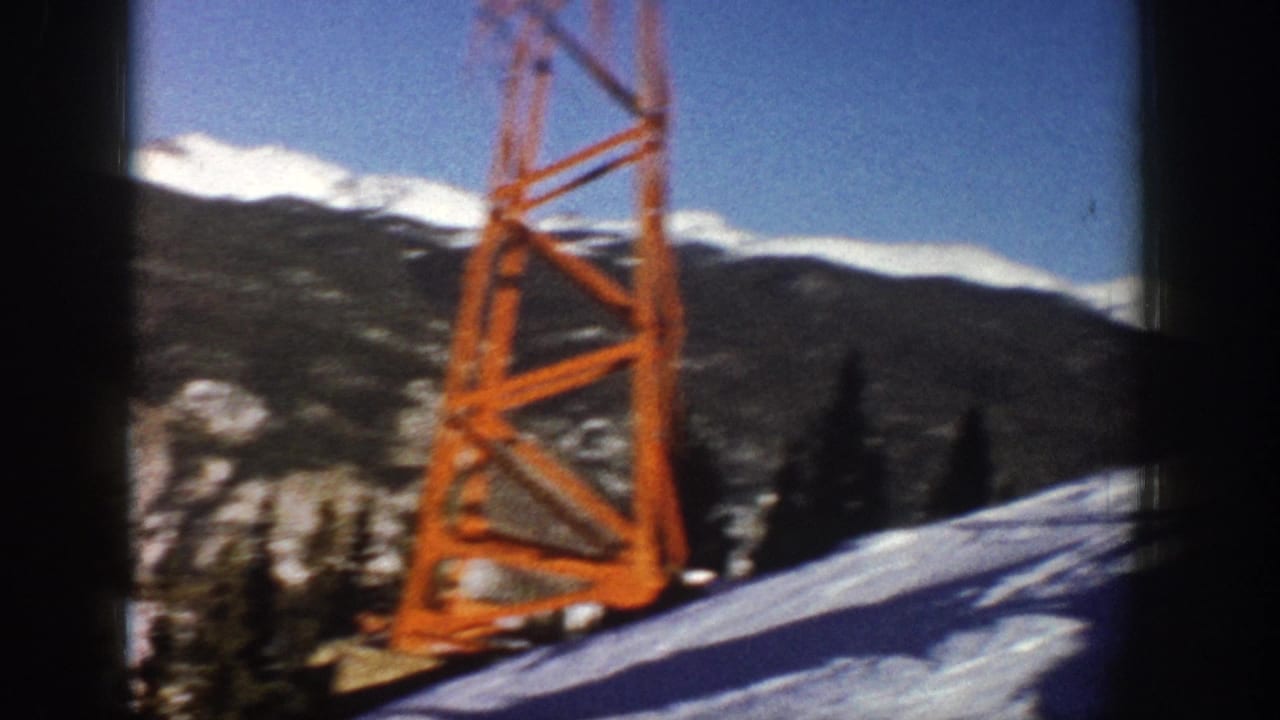 1961 Aspen home-movie frame showing an orange ski lift tower on snow with dark mountain ridges and white peaks in the background.