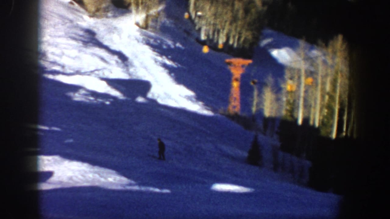 1961 Aspen frame with a distant skier descending a snowy run beside trees and orange lift infrastructure.