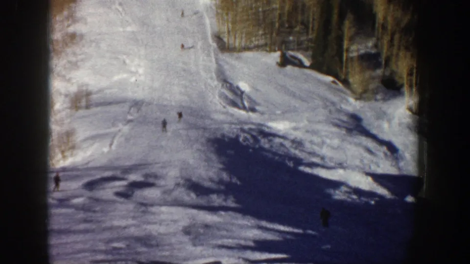 Wide 1961 Aspen ski slope view with several distant skiers descending a sunlit run bordered by trees.