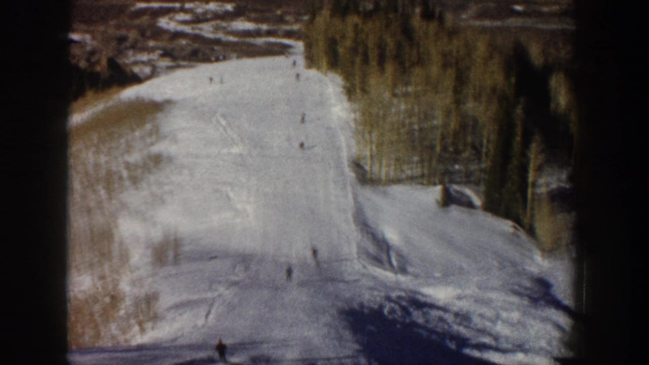 Archival Aspen ski footage frame showing distant skiers on a snowy hillside with trees lining the upper edge.
