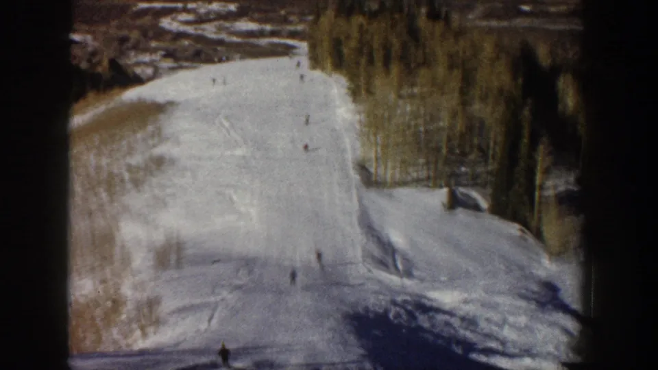 Archival Aspen ski footage frame showing distant skiers on a snowy hillside with trees lining the upper edge.