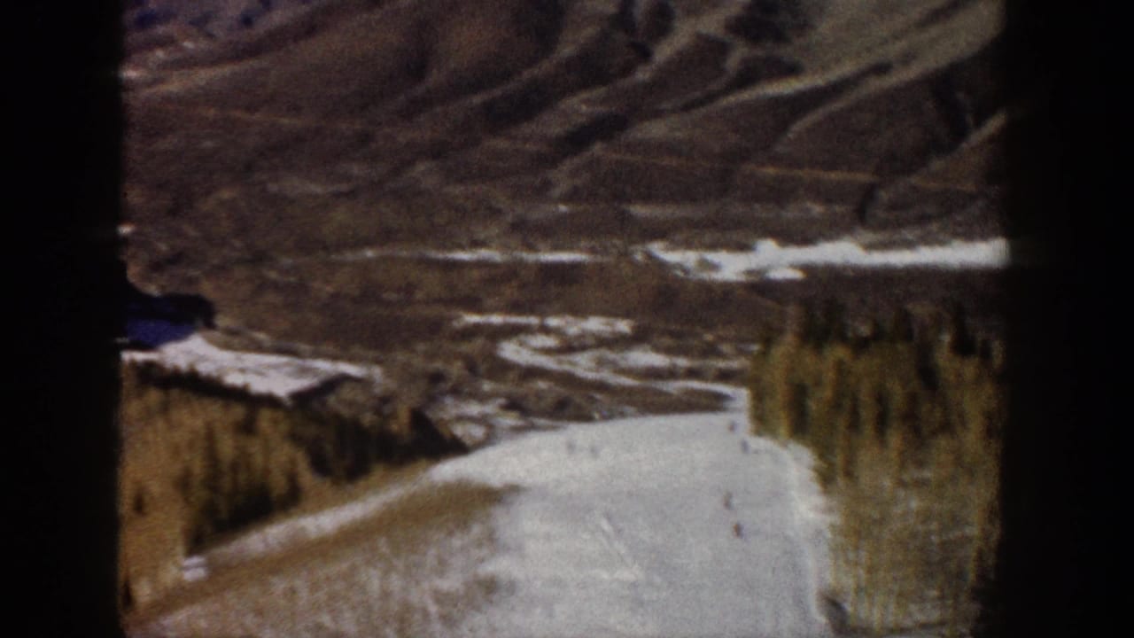 Aspen winter home-movie frame showing a snow run below darker mountain terrain, with a small skier visible near the slope.