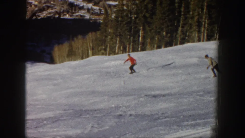 1961 Aspen ski-trip frame showing two distant skiers, including one in red, descending a snowy slope near trees.