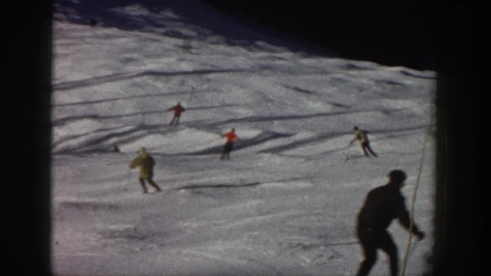 Vintage 8mm frame of recreational skiers descending an Aspen slope, with a dark foreground figure entering at right.