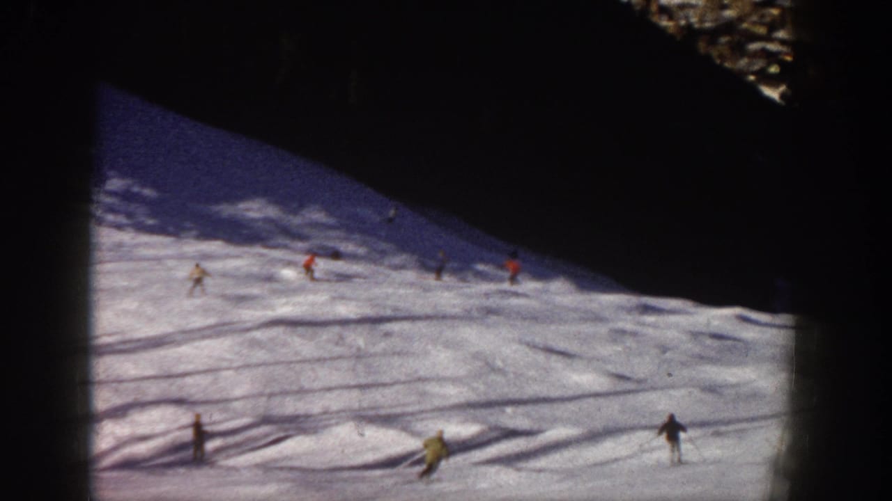 1961 Aspen home-movie frame showing several skiers spread across a snowy slope under dark mountain shadow.