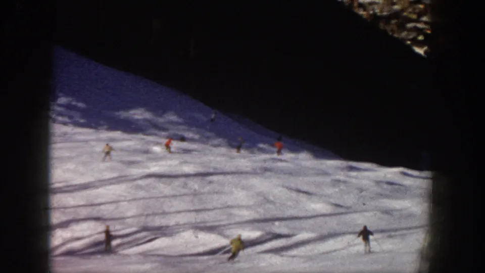 1961 Aspen home-movie frame showing several skiers spread across a snowy slope under dark mountain shadow.