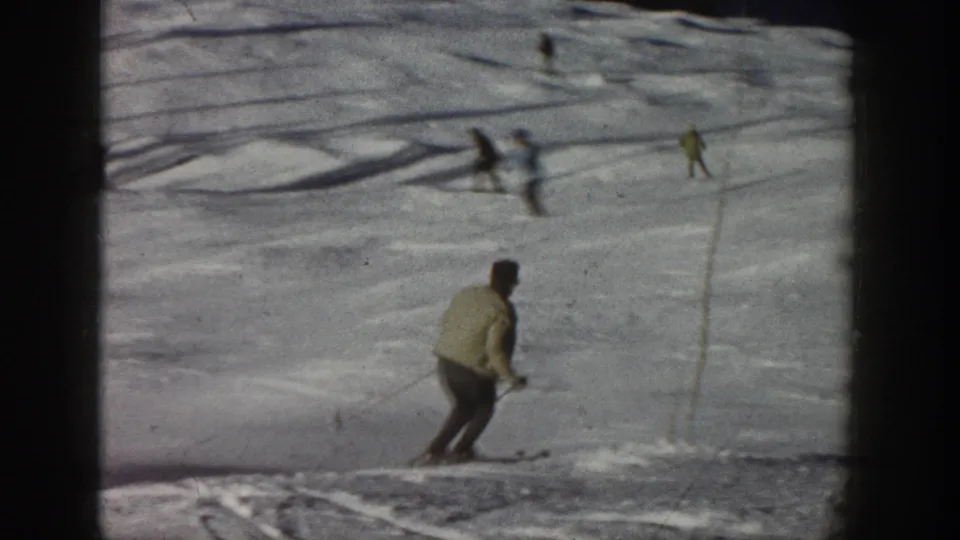 1961 Aspen home-movie view of a skier near the lower edge of the frame, with poles or markers and distant skiers on the snow.