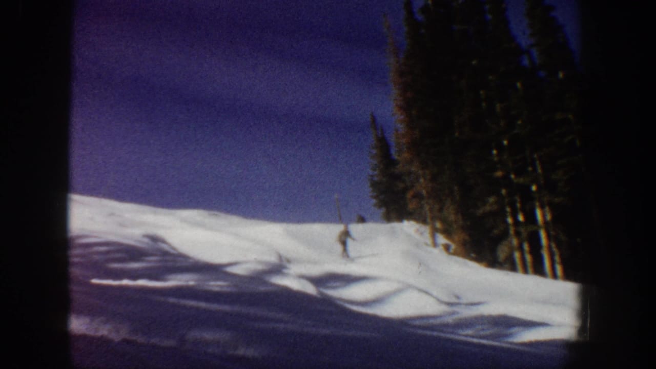 1961 Aspen ski footage frame showing a distant skier turning across snow near a stand of tall pines.