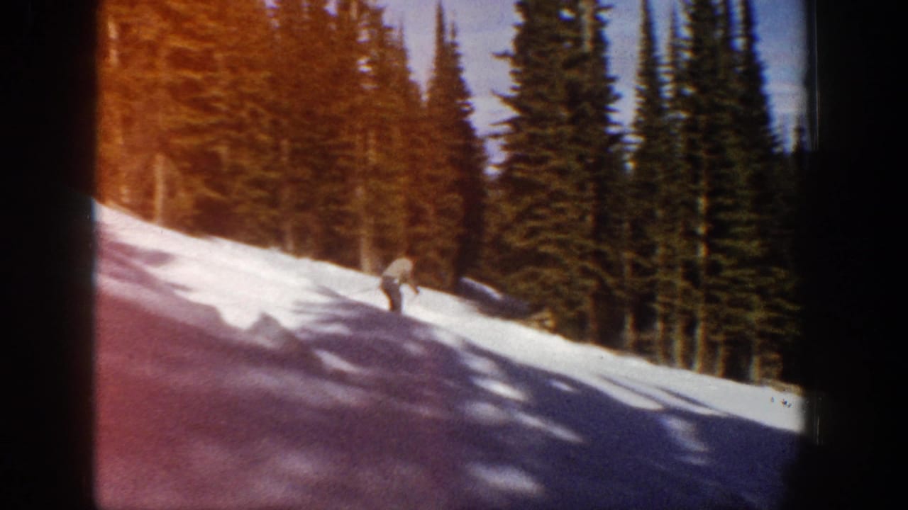 Aspen 1961 home-movie frame showing a skier low on the snowy slope, with dense evergreens and reddish film flare along the edge.