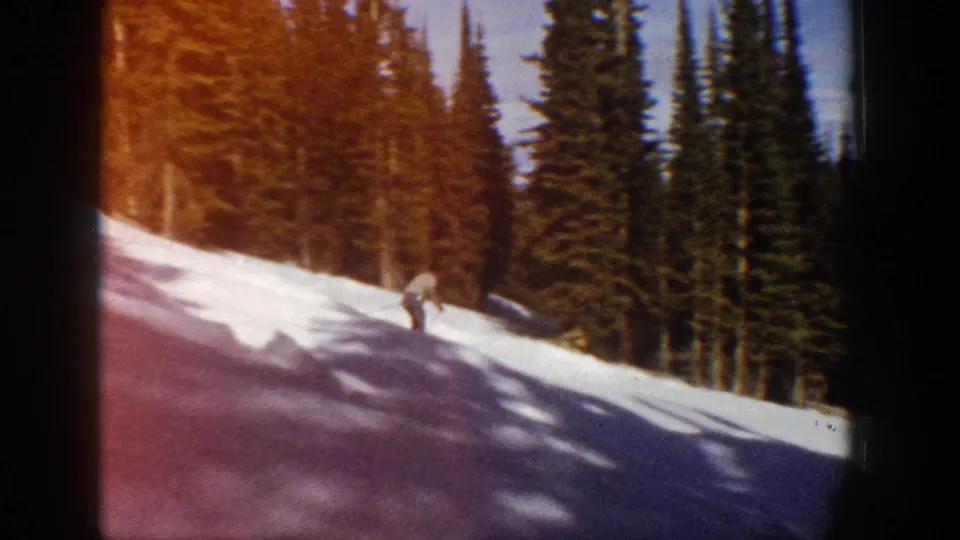 Aspen 1961 home-movie frame showing a skier low on the snowy slope, with dense evergreens and reddish film flare along the edge.