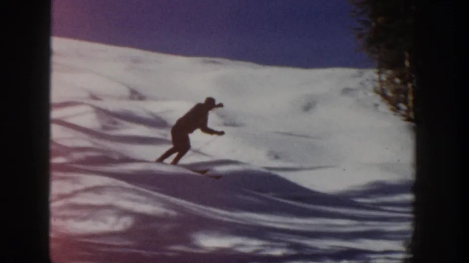 The lone skier leans into the descent on an Aspen snow slope, with evergreen trees and dark film edges visible.