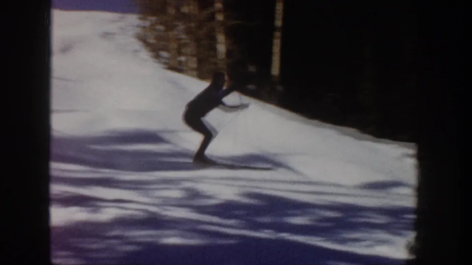 A skier in dark clothing moves across a snowy Aspen slope in 1961, bordered by evergreen trees and long winter shadows.