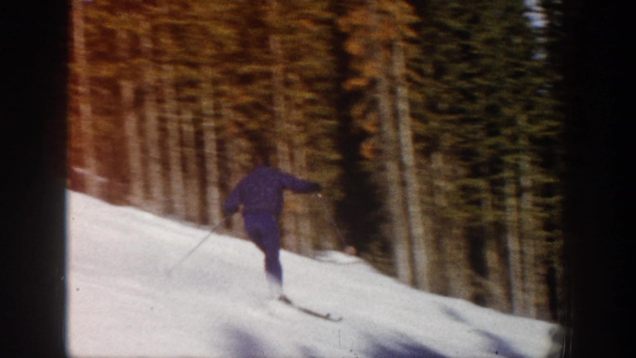 The skier is seen from behind on a snowy Aspen run, moving away toward a stand of tall trees in the 1961 home-movie footage.