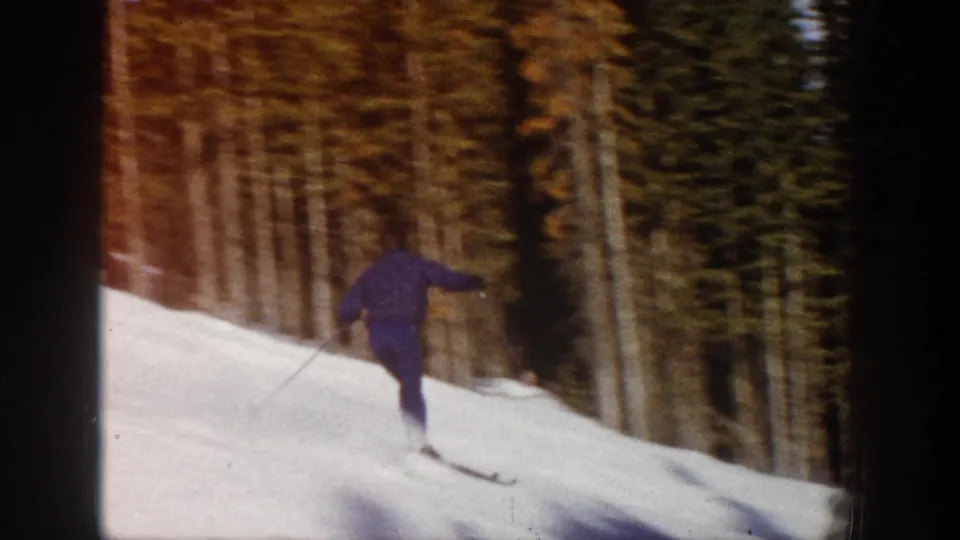 The skier is seen from behind on a snowy Aspen run, moving away toward a stand of tall trees in the 1961 home-movie footage.